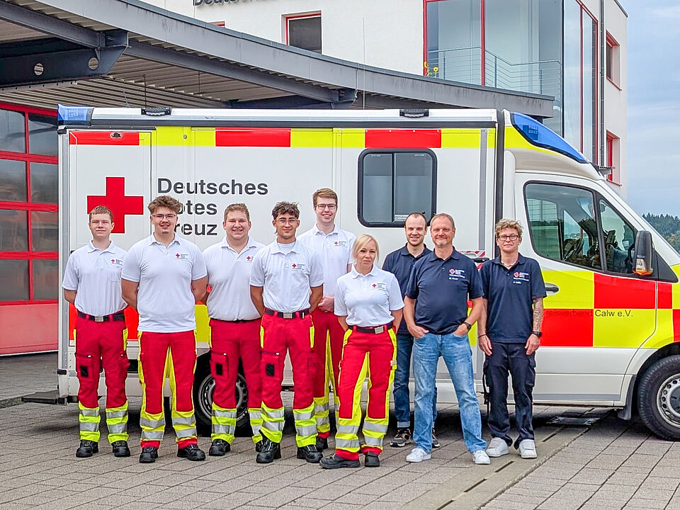 2026-10-01_Azubis Sechs junge Menschen in Rettungsdienstkleidung mit ihren Ausbildern vor einem Rettungswagen.